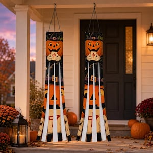 May include: Two hanging windsock decorations featuring a graphic of a smiling jack-o'-lantern wearing a witch hat and holding a mahjong tile with the Chinese character for middle. The fabric is black with white vertical streamers at the bottom.