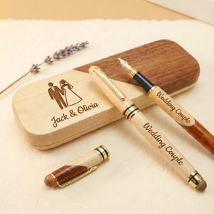 May include: A wooden fountain pen and a matching wooden ballpoint pen with a stylus tip, both engraved with the text Wedding Couple. They are accompanied by a rectangular wooden case featuring an engraved illustration of a couple and the text Jack & Olivia.