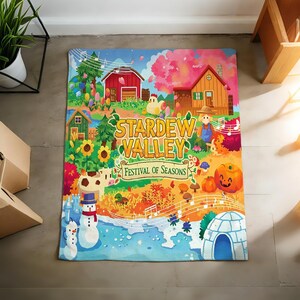 Pu&ograve; includere: Una coperta colorata con la scritta "Stardew Valley Festival of Seasons". Il design include una scena di fattoria con un fienile rosso, una casa, girasoli, pupazzi di neve e una zucca. La coperta ha uno stile vivace e fumettistico.