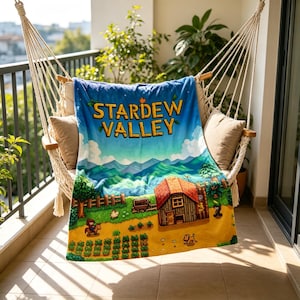 Pu&ograve; includere: Una coperta blu con la scritta "STARDEW VALLEY" in giallo. La coperta presenta un paesaggio colorato con una casa, una fattoria e montagne. &Egrave; drappeggiata su un'amaca.