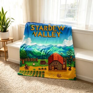 Pu&ograve; includere: Una morbida coperta con il logo del gioco "Stardew Valley" e un paesaggio colorato. Il design include una casa con il tetto rosso, campi, alberi e montagne sotto un cielo blu. La coperta &egrave; piegata su una panca bianca vicino a una finestra.
