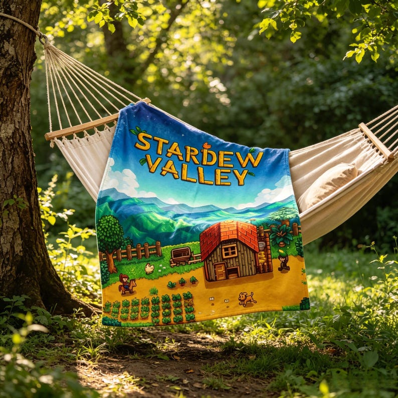 Pu&ograve; includere: Una coperta di Stardew Valley &egrave; appoggiata su un'amaca. La coperta presenta un paesaggio colorato con il logo del gioco, una casa e un contadino. La scena &egrave; ambientata in un ambiente naturale con alberi e fogliame verdi.