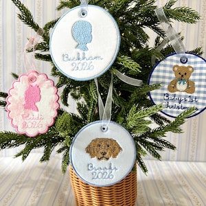 May include: Four circular and scalloped embroidered ornaments hanging on a small tree. One features a blue silhouette with "Beckham 2026", one a pink silhouette with "Reidi 2026", one a brown dog with "Brooks 2026", and one a teddy bear with "Baby's 1st Christmas".