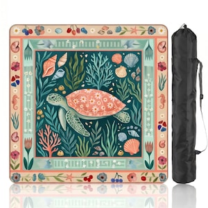 May include: A square beach mat featuring a sea turtle design with coral, seashells, and aquatic plants. The turtle has a pink shell with a floral pattern. The mat is surrounded by a decorative border and comes with a black carrying bag.