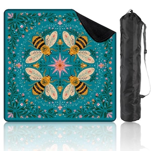 May include: A teal picnic blanket with a bee and floral design. The blanket features four large bees with yellow and black stripes, surrounded by pink and white flowers. A black carrying bag is included.