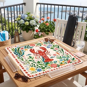 Puede incluir: Una mesa de madera con una alfombra decorativa con un dise&ntilde;o de langosta roja y vida marina. La alfombra est&aacute; rodeada de bandejas de pl&aacute;stico transparente. Gafas de sol y una bebida est&aacute;n sobre la mesa, con flores en macetas y vista al agua al fondo.