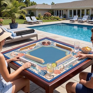 Op de afbeelding: Een vierkante houten Mahjong-tafel met een blauw speeloppervlak en een rand met marinethema. De tafel is opgesteld voor een spel, met tegels op het oppervlak. De tafel staat buiten bij een zwembad.