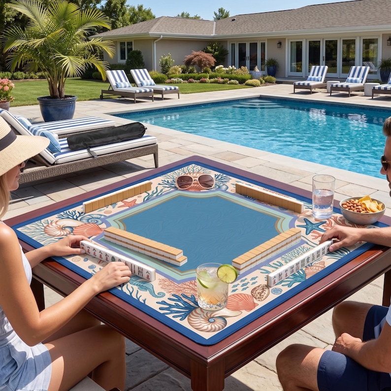 Op de afbeelding: Een vierkante houten Mahjong-tafel met een blauw speeloppervlak en een rand met marinethema. De tafel is opgesteld voor een spel, met tegels op het oppervlak. De tafel staat buiten bij een zwembad.
