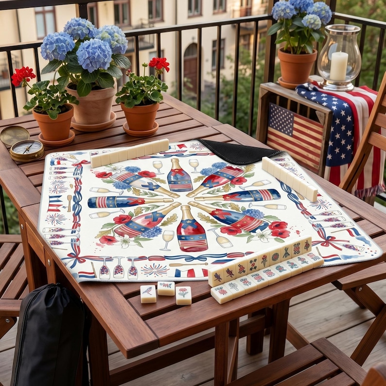K&ouml;nnte beinhalten: Ein Holztisch, der f&uuml;r ein Mahjong-Spiel gedeckt ist, mit einer dekorativen Spielunterlage mit einem patriotischen Design aus Champagnerflaschen, Blumen und Feuerwerk. Der Tisch ist von Topfpflanzen und einer dekorativen amerikanischen Flagge umgeben.