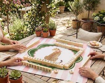May include: A mahjong set featuring ivory-colored tiles with black and red characters, arranged on a square play mat. The mat has a pink and white striped border and a central illustration of a leopard wearing a pink bow.