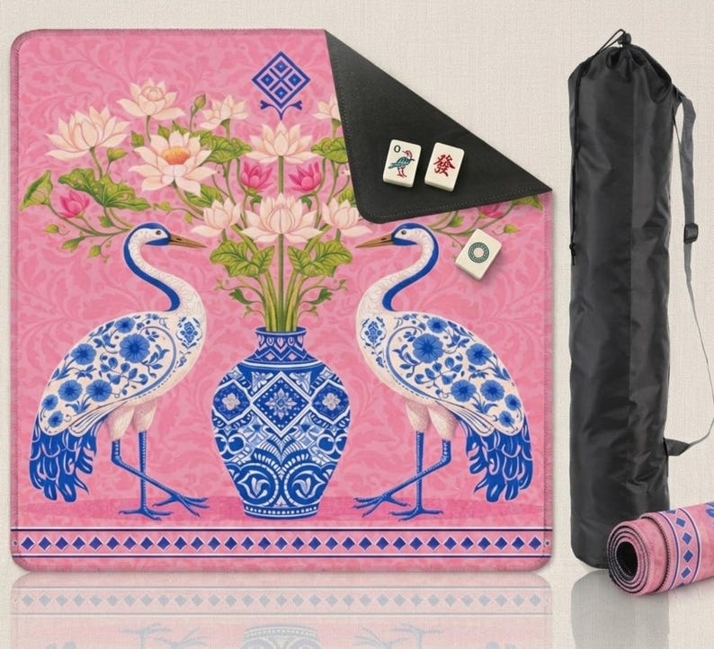 May include: A pink yoga mat with a floral and crane design. The mat features a blue and white vase with flowers, two cranes, and a decorative border. A black carrying bag and a rolled-up mat are also visible.