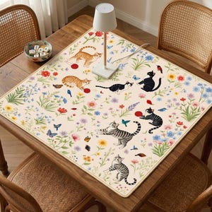 May include: A rectangular beige desk mat featuring a floral and cat design. The mat has illustrations of various cats, flowers, butterflies, and birds. The mat is placed on a wooden table with a lamp and a small bowl.