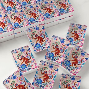 May include: A set of rectangular mahjong tiles featuring a repeating illustration of a brown monkey holding a green branch, surrounded by blue lotus flowers and pink floral accents on a light beige background with white sides.