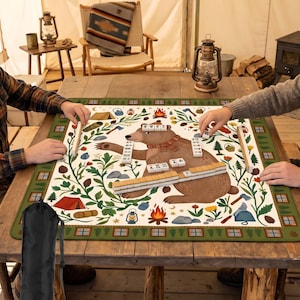 May include: A Mahjong game mat with a bear illustration, surrounded by camping-themed icons. The mat is green with a white center, and the bear is brown. The game is set up on a rustic wooden table, with players' hands visible.