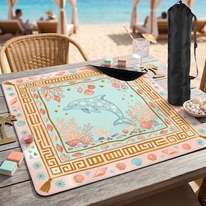 Op de afbeelding: Een strandlaken met een dolfijnontwerp. Het laken heeft een lichtblauwe achtergrond met illustraties van dolfijnen, koraal en schelpen. De rand is een mix van roze, goud en blauw. Een zwarte draagtas is inbegrepen.
