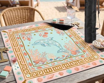 May include: A beach blanket with a dolphin design. The blanket features a light blue background with a dolphin, coral, and seashell illustrations. The border is a mix of pink, gold, and blue. A black carrying bag is included.