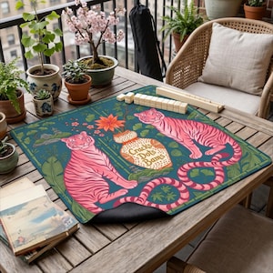 May include: A teal Mahjong mat with two pink tigers and floral details. The mat has the text "Cracks Dots Bams" on a vase. Mahjong tiles and racks are visible. The scene is set on a wooden table.