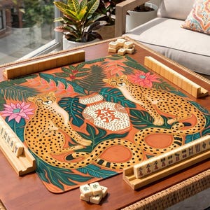 May include: A wooden mahjong table with a colorful mat featuring cheetahs and tropical foliage. The table is set with wooden tile racks and tiles, ready for play. The table is on a patio with a view of a building.