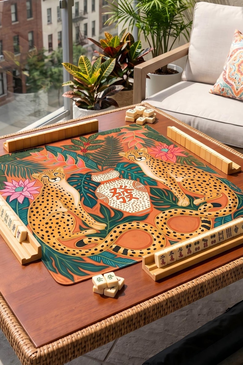 May include: A wooden mahjong table with a colorful mat featuring cheetahs and tropical foliage. The table is set with wooden tile racks and tiles, ready for play. The table is on a patio with a view of a building.