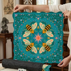 Puede incluir: Una esterilla cuadrada turquesa con borde festoneado, con un diseño simétrico de cuatro abejas, flores y una estrella. La esterilla está enrollada en una funda de transporte negra. La esterilla es probablemente para un juego.