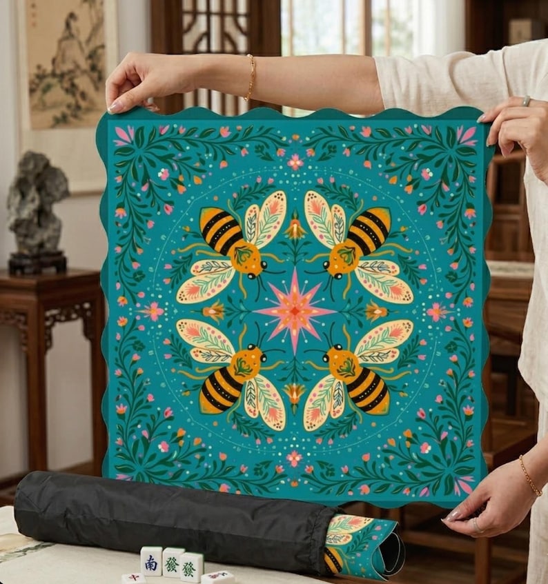 May include: A teal square mat with a scalloped edge, featuring a symmetrical design of four bees, flowers, and a starburst. The mat is rolled up in a black carrying case. The mat is likely for a game.