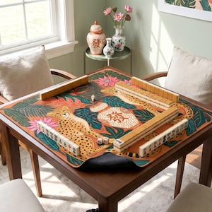 May include: A square wooden mahjong table with a colorful mat featuring cheetahs and floral designs. The table is set with tiles and dice, ready for play. The room has a window and decorative vases.