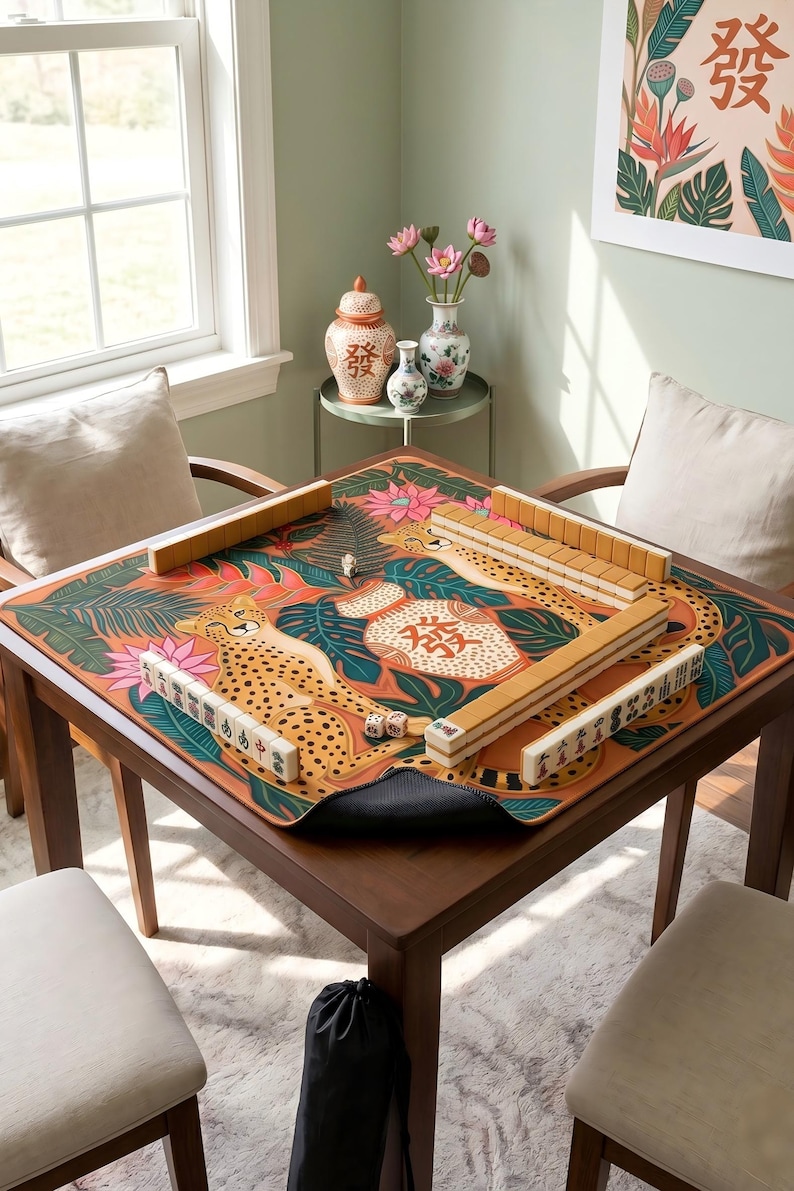 May include: A square wooden mahjong table with a colorful mat featuring cheetahs and floral designs. The table is set with tiles and dice, ready for play. The room has a window and decorative vases.