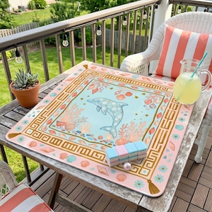 May include: A square, pink and blue mat with a dolphin illustration, seashells, and coral. The mat has a gold border with a Greek key design. A pitcher of lemonade and a set of colorful blocks sit on the mat, which is on a wooden table.