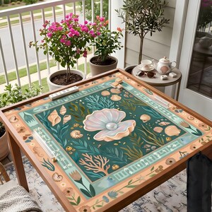 Puede incluir: Una mesa de madera con un dise&ntilde;o marino. La mesa tiene un fondo verde azulado con ilustraciones de conchas marinas, corales y una gran perla dentro de una ostra abierta. La mesa est&aacute; rodeada de plantas en macetas y un balc&oacute;n.