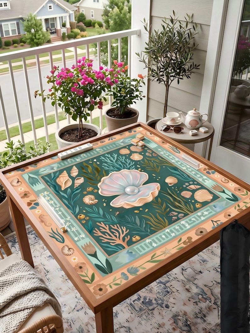 Puede incluir: Una mesa de madera con un dise&ntilde;o marino. La mesa tiene un fondo verde azulado con ilustraciones de conchas marinas, corales y una gran perla dentro de una ostra abierta. La mesa est&aacute; rodeada de plantas en macetas y un balc&oacute;n.