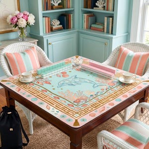 May include: A square wooden table with a dolphin-themed game mat, surrounded by white wicker chairs with striped pillows. The mat features a dolphin, coral, and seashell design in pastel colors. Game pieces and teacups are on the table.