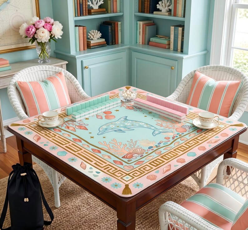 May include: A square wooden table with a dolphin-themed game mat, surrounded by white wicker chairs with striped pillows. The mat features a dolphin, coral, and seashell design in pastel colors. Game pieces and teacups are on the table.