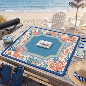 Op de afbeelding: Een vierkante mahjong-speelmat met een strandthema. De mat heeft een blauw midden, een rand met koraal, schelpen en zeepaardjes en een donkerblauwe rand. Mahjong-tegels liggen op de mat. De sc&egrave;ne speelt zich af op een houten tafel bij het strand.