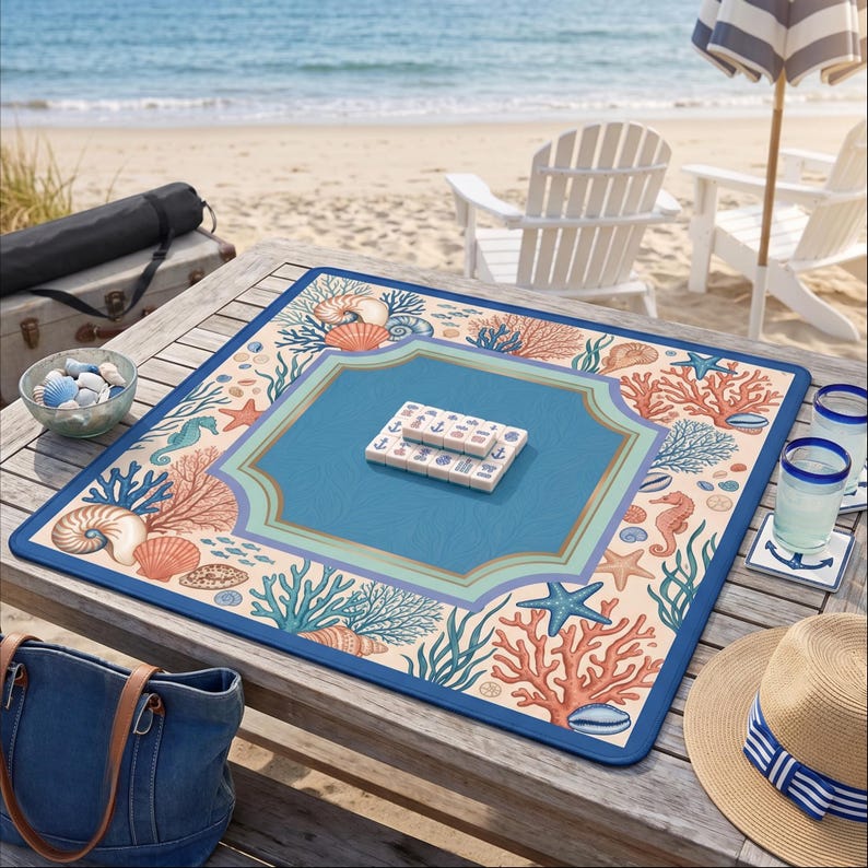 Op de afbeelding: Een vierkante mahjong-speelmat met een strandthema. De mat heeft een blauw midden, een rand met koraal, schelpen en zeepaardjes en een donkerblauwe rand. Mahjong-tegels liggen op de mat. De sc&egrave;ne speelt zich af op een houten tafel bij het strand.