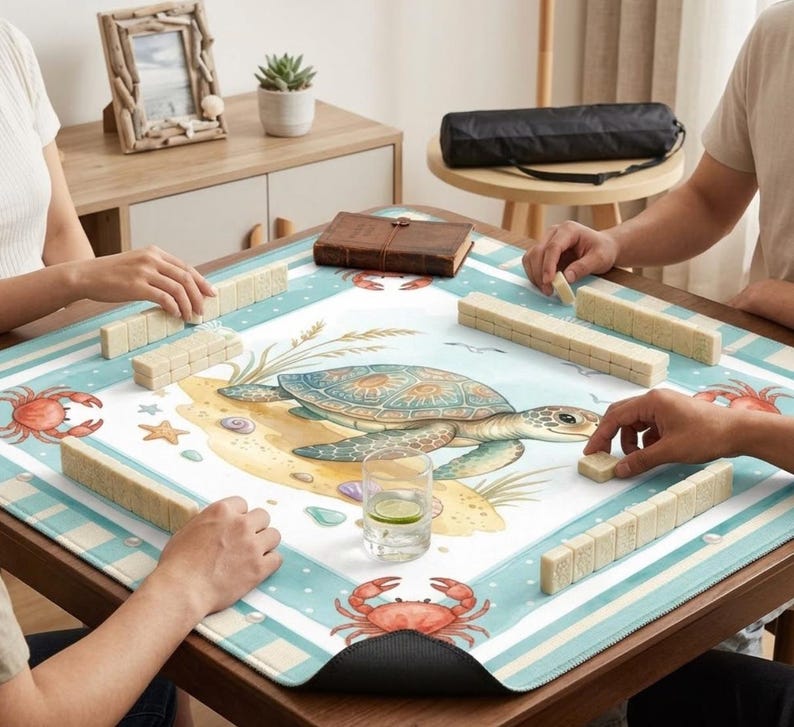 May include: A square game table with a sea turtle design. The table is covered with a blue and white mat featuring a turtle, crabs, and seashells. Players are arranging beige game tiles. A glass of water with a lemon slice is on the table.