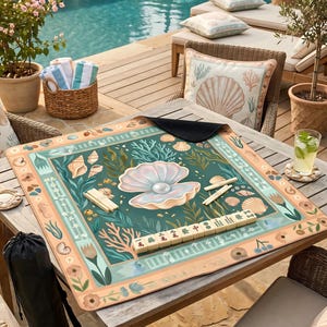 May include: A square mahjong game mat with a sea-themed design featuring a large pearl, seashells, and coral in shades of teal, peach, and cream. The mat is set on a wooden table outdoors near a pool.