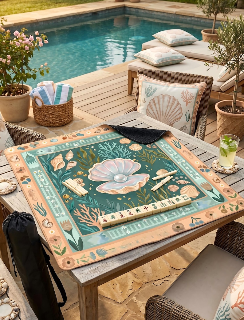 Puede incluir: Tapete de juego de mahjong cuadrado con un dise&ntilde;o tem&aacute;tico marino que presenta una gran perla, conchas marinas y coral en tonos de verde azulado, melocot&oacute;n y crema. El tapete est&aacute; colocado sobre una mesa de madera al aire libre, cerca de una piscina.