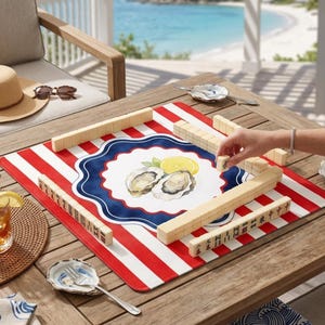May include: A red and white striped Mahjong game mat with a central design of oysters and lemons. Wooden Mahjong tiles are arranged on the mat. A person's hand is placing a tile. The scene is set on a wooden table.