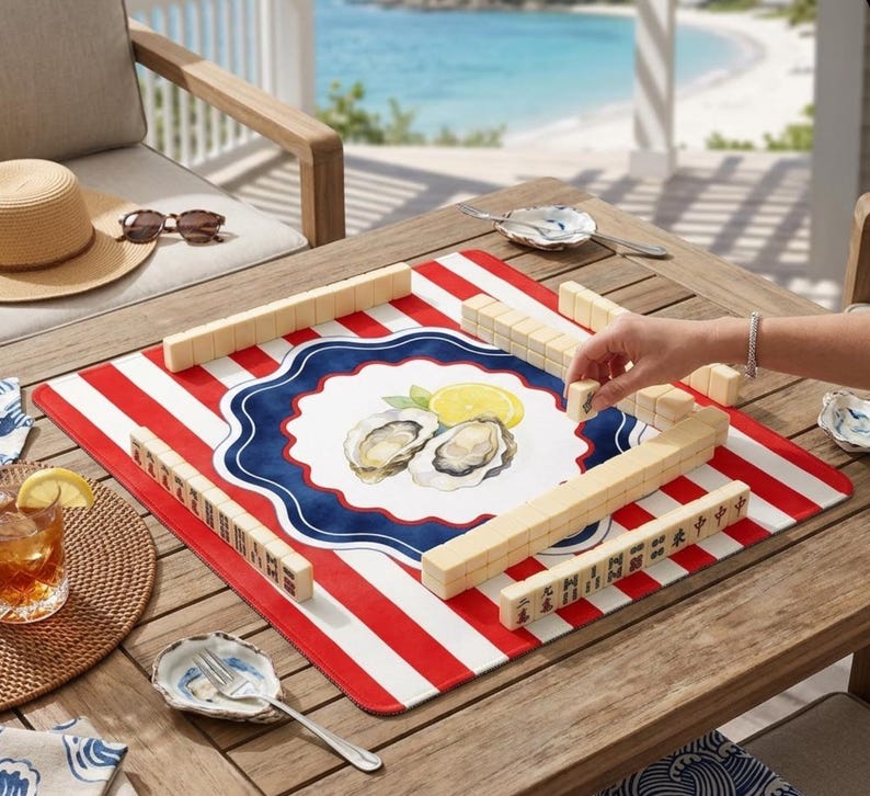 May include: A red and white striped Mahjong game mat with a central design of oysters and lemons. Wooden Mahjong tiles are arranged on the mat. A person's hand is placing a tile. The scene is set on a wooden table.