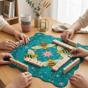 May include: A teal Mahjong game set with a floral and bee design. The game is set up on a teal mat with a floral and bee pattern. The wooden tiles are being arranged by several people. A stack of ceramic cups and a candle holder are in the background.