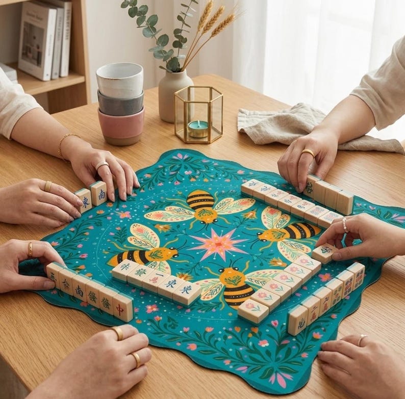 May include: A teal Mahjong game set with a floral and bee design. The game is set up on a teal mat with a floral and bee pattern. The wooden tiles are being arranged by several people. A stack of ceramic cups and a candle holder are in the background.