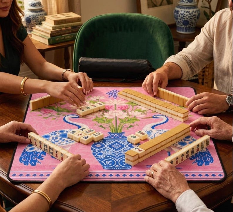 May include: A group of people playing Mahjong. The game is set on a pink mat with a floral and bird design. The tiles are beige and the table is a dark wood.