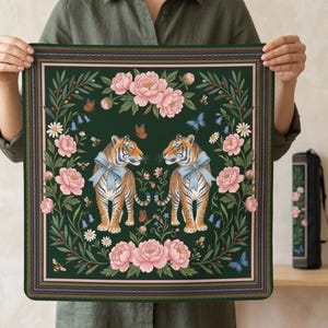 Könnte beinhalten: Quadratisches dekoratives Textil mit dunkelgrünem Hintergrund mit zwei Tigern mit blauen Schleifen, umgeben von rosa Pfingstrosen, Schmetterlingen und Blättern. Der Rand hat ein rosa und grünes Design.