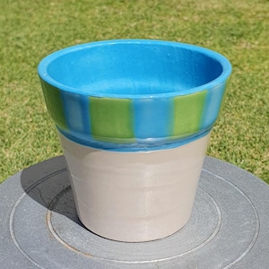 May include: A ceramic plant pot with a light beige base and a blue and green striped rim. The pot is on a grey pottery wheel. The background features green grass and a textured wall.