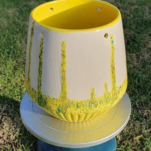 May include: A ceramic planter with a yellow interior and exterior rim. The off-white exterior features green and yellow painted accents. The planter sits on a silver and blue stand, with a grassy background.