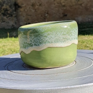May include: A handmade ceramic bowl with a green base and a white and green rim. The bowl sits on a grey pottery wheel, which is on a blue stand, against a backdrop of green grass and a wall.