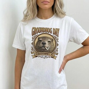 Puede incluir: Camiseta blanca con un diseño del Día de la Marmota. El diseño presenta una marmota fumando un cigarrillo, con las palabras "GROUNDHOG DAY" y "PUNXSUTAWNEY, PA EST 1887". El diseño es en tonos marrones y dorados.