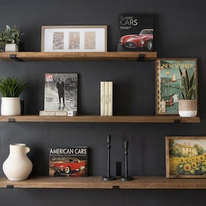 May include: Three wooden floating shelves with black metal brackets are mounted on a dark gray wall. The shelves display various decorative items, including potted plants, framed art, magazines, and a white vase. The top shelf holds a framed picture and a magazine with a red car on the cover.