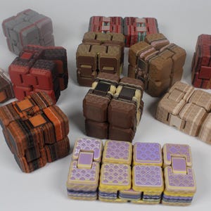May include: A selection of wooden fidget toys in different colours and patterns. The toys are cubes and rectangular shapes, with colours including brown, red, grey, and yellow. Some have geometric designs, others are solid.