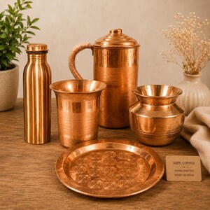Puede incluir: Una colección de utensilios de cocina de cobre hechos a mano, que incluyen una botella de agua, un vaso, una jarra con asa trenzada, una olla pequeña y un plato decorativo. El plato tiene un patrón circular. Los artículos se exhiben sobre una superficie de madera.